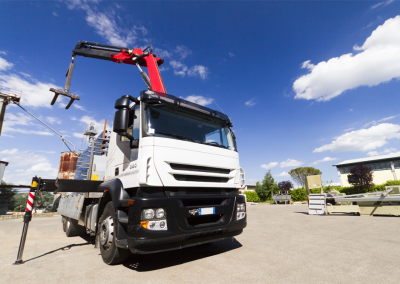 A white flatbed truck with a red crane arm is parked outdoors on a sunny day, lifting construction materials. The scene, ideal for crane audits, features a blue sky with scattered clouds and buildings visible in the background.