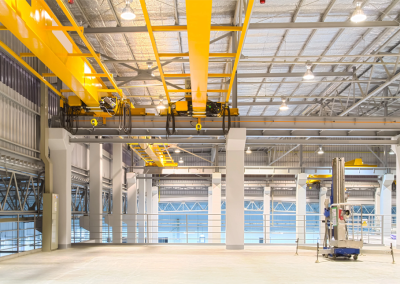 Interior of a brightly lit industrial facility featuring yellow overhead cranes, metal beams, and a scissor lift positioned on a clean, spacious concrete floor—ideal for crane audits and equipment inspections.