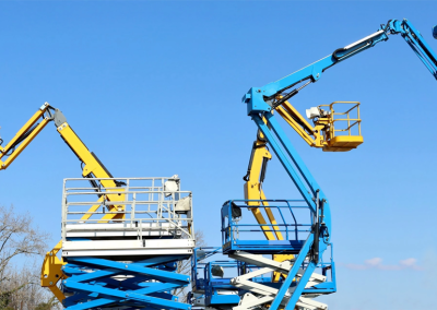 Several yellow and blue articulated boom lifts with workers in safety gear extend upward against a clear blue sky, illustrating the importance of crane audits. Some trees are visible in the background.