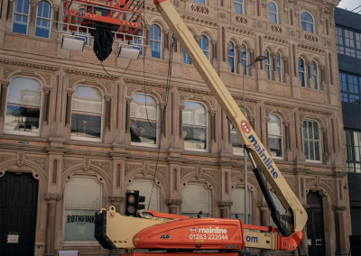 A cherry picker lift with a worker platform is raised beside a tall, ornate building. Traffic cones surround the vehicle, and black fabric or cables hang from the platform above the street during crane audits.