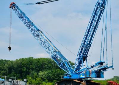 A large blue crawler crane with an extended boom stands on a paved lot, ideal for crane audits, surrounded by trees and machinery under a partly cloudy sky.