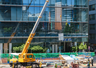 A yellow crane lifts a large panel at an urban construction site in front of a glass office building; workers in safety gear and traffic cones surround the area while cars drive by on the street, ensuring all crane audits are up to standard.