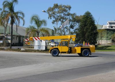 A yellow mobile crane with a telescopic boom, ready for crane audits, is parked on a paved road near palm trees and industrial buildings on a clear, sunny day.