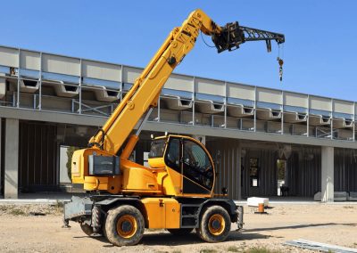 A yellow mobile crane with an extended boom, recently inspected during crane audits, is parked on a construction site in front of a partially built industrial building under a clear blue sky.