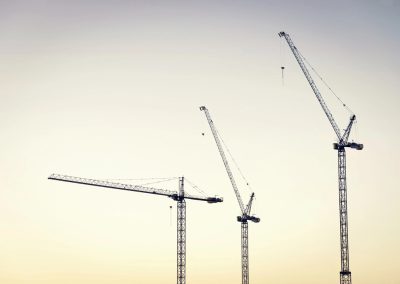 Three tall construction cranes stand against a pale sky at sunset or sunrise, with no buildings visible—an industrial scene that echoes the precision and order of crane audits in a minimalist landscape.