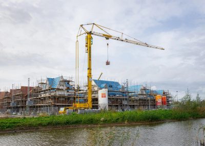 A construction site with houses covered in scaffolding and blue roofing, a tall yellow crane undergoing crane audits while lifting materials, and a pond with green vegetation in the foreground under a cloudy sky.