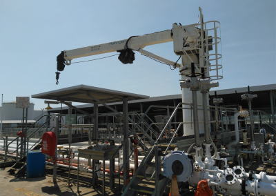 A white industrial crane with a hook is mounted on a platform surrounded by pipes, valves, and metal structures at an outdoor industrial facility under a clear sky—an ideal setting for thorough crane audits.